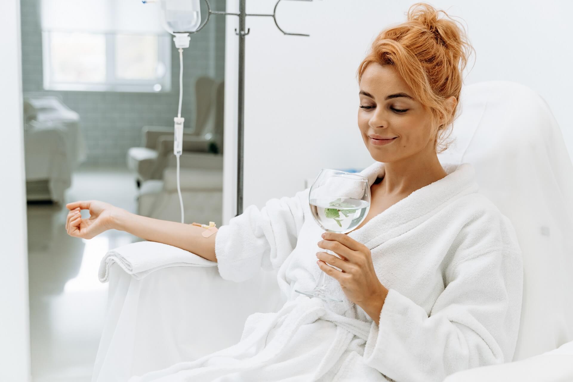 Side view portrait of charming woman in white bathrobe sitting in armchair and holding glass of lemon water. | Estevez Aesthetics in Las Vegas, NV