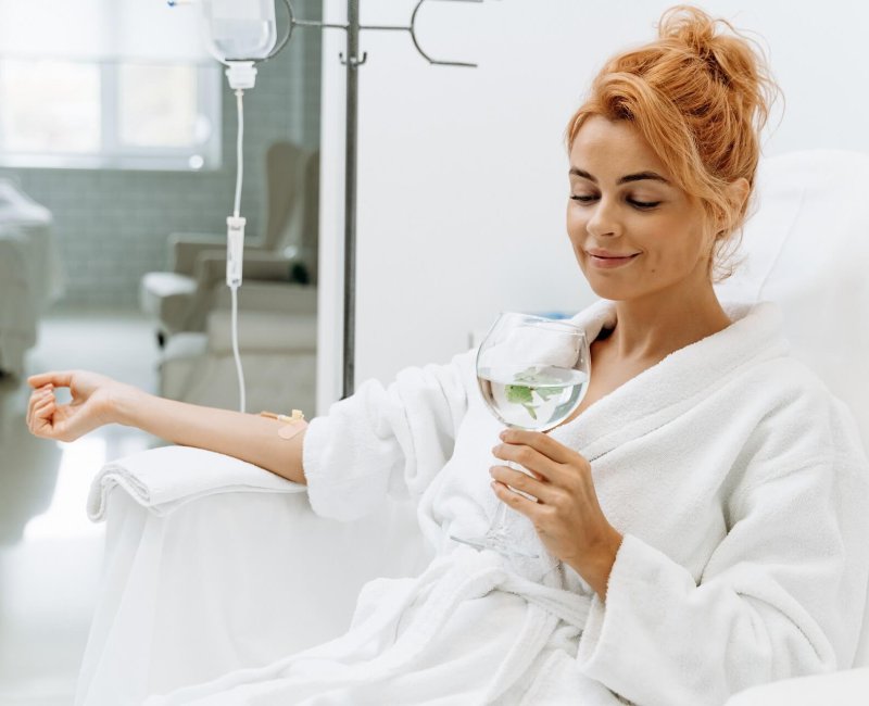 Side view portrait of charming woman in white bathrobe sitting in armchair and holding glass of lemon water. | Estevez Aesthetics in Las Vegas, NV